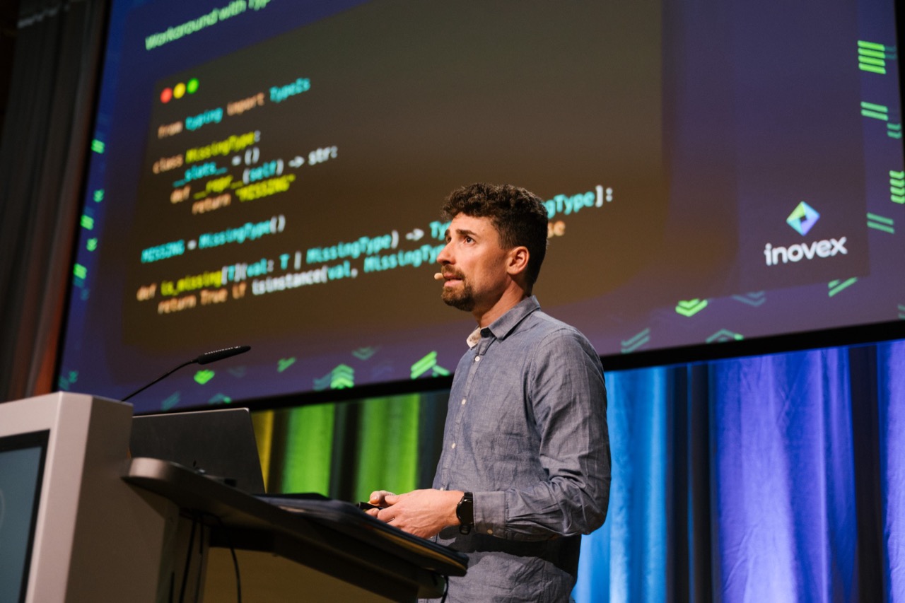 A speaker at the lectern in front of a large screen showing Python code